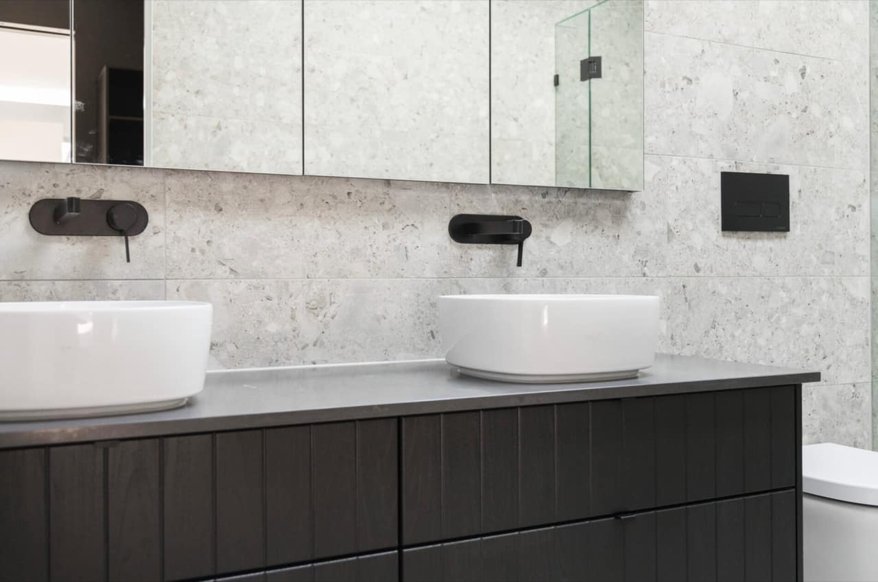Black-stained timber vanity with grey stone top, twin round white basins and matte black tapware on terrazzo-look tiles