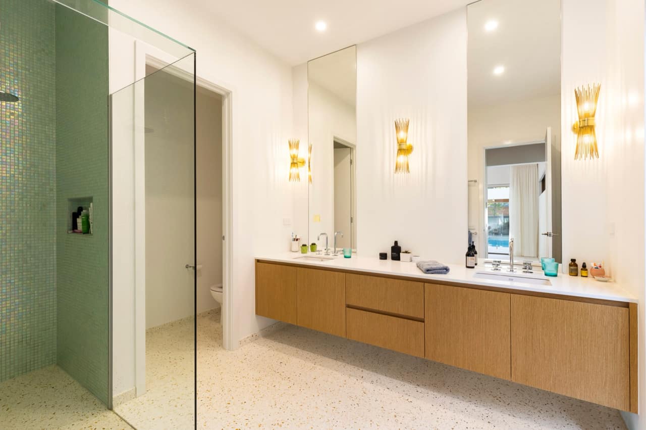 Long oak floating vanity with twin undermount basins, brass sconces and terrazzo floor