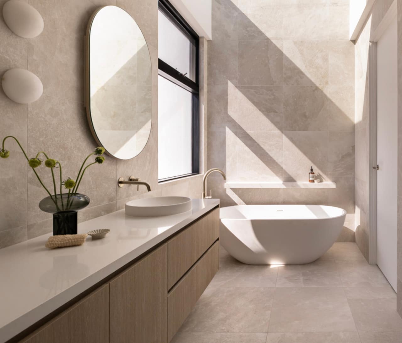 Sunlit limestone bathroom with light oak floating vanity, oval mirror and freestanding bath