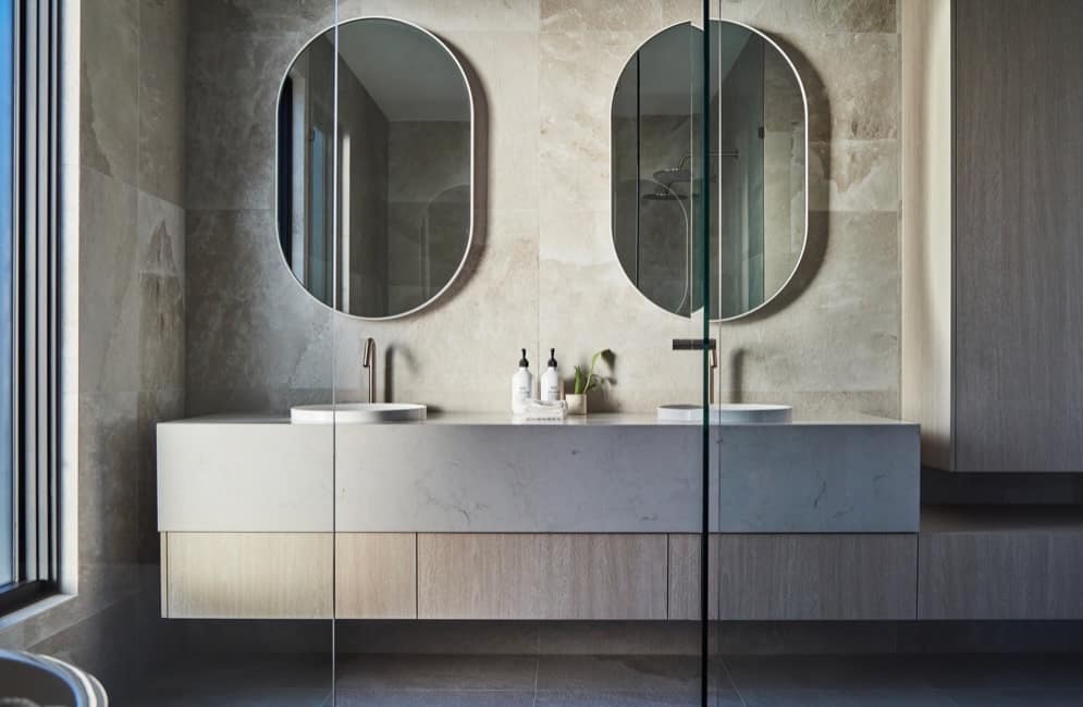 Stone-wrapped floating vanity with twin basins and oval mirrors behind a glass shower screen