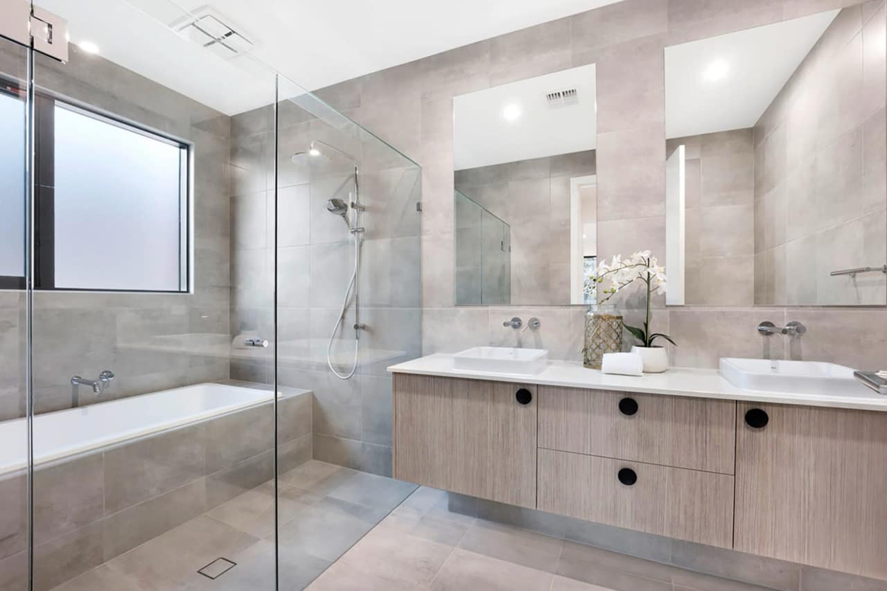 Washed-oak floating vanity with twin square basins and round black fingerpulls beside a frameless glass shower